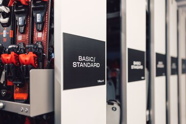 Mehrere Skidepots im Sportverleih sind mit schwarzen Schildern beschriftet, auf denen „Basic / Standard“ steht; links sind eingesetzte Skier mit Bindungen sichtbar. | © Gargellner Bergbahnen GmbH & Co KG, Daniel Zangerl