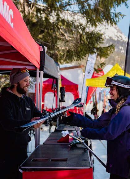 Eine Frau in Skikleidung mit Helm und Rucksack steht an einem Teststand im Freien und bekommt von einem Mitarbeiter unter einem roten Zelt ein Paar Ski überreicht. Im Hintergrund sind weitere bunte Zelte, Banner und verschneite Berge zu sehen. | © Montafon Tourismus GmbH, Marie Rudigier