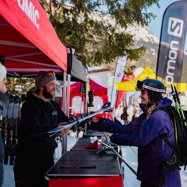 Eine Frau in Skikleidung mit Helm und Rucksack steht an einem Teststand im Freien und bekommt von einem Mitarbeiter unter einem roten Zelt ein Paar Ski überreicht. Im Hintergrund sind weitere bunte Zelte, Banner und verschneite Berge zu sehen. | © Montafon Tourismus GmbH, Marie Rudigier