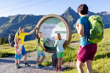 Eine funfköpfige Familie die am Erlebnisberg Golm auf Golmis Forschungspfad wandert und auf eine runde Tafel blickt auf der steht, wie man Golm Forscher wird | © Golm Silvretta Lünersee Tourismus GmbH Bregenz, Stefan Kothner